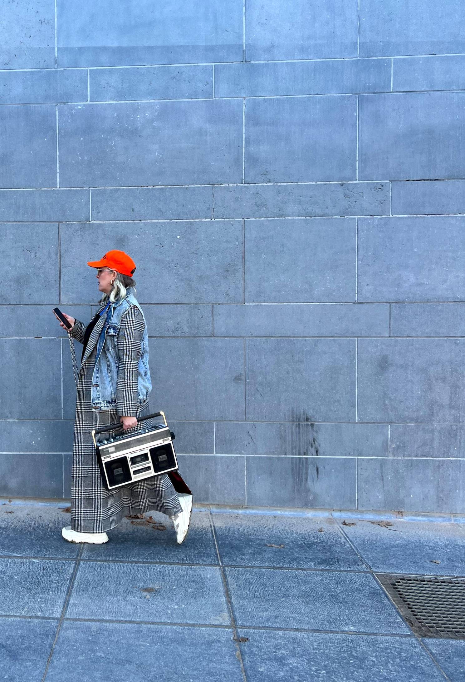 Jessie in an orange cap and checked trousers walking past a grey wall with a boombox