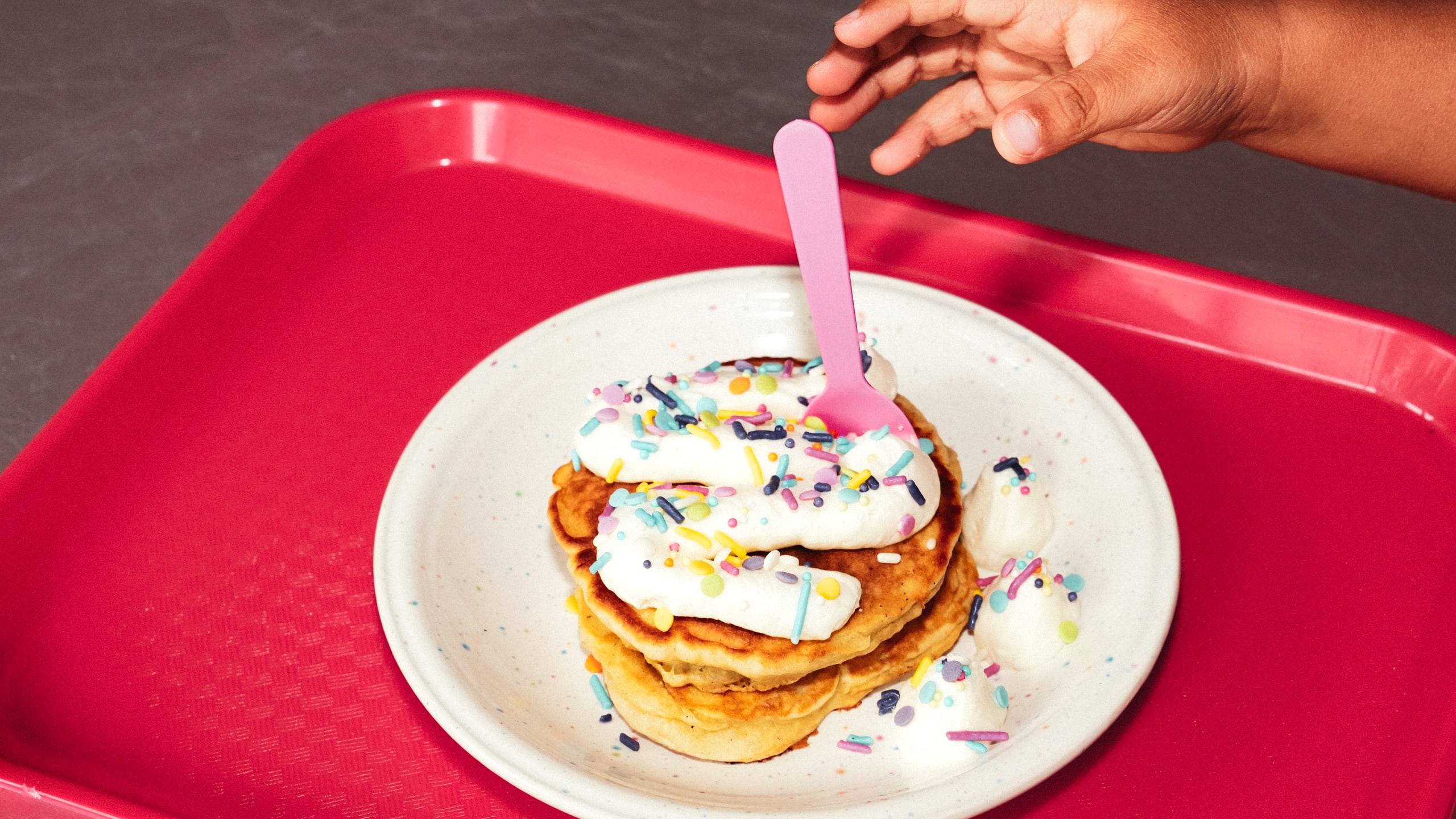 A kid reaching for a pancake stack with whipped cream and sprinkles on a red tray