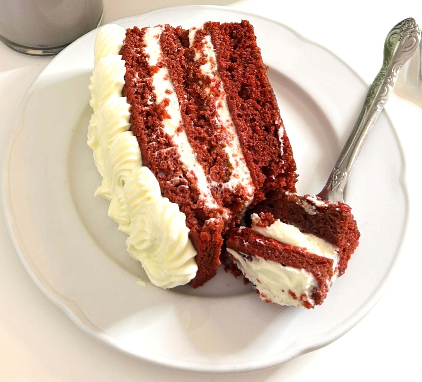 A thick slice of red velvet cake with cream cheese frosting between the layers, a fork on the side